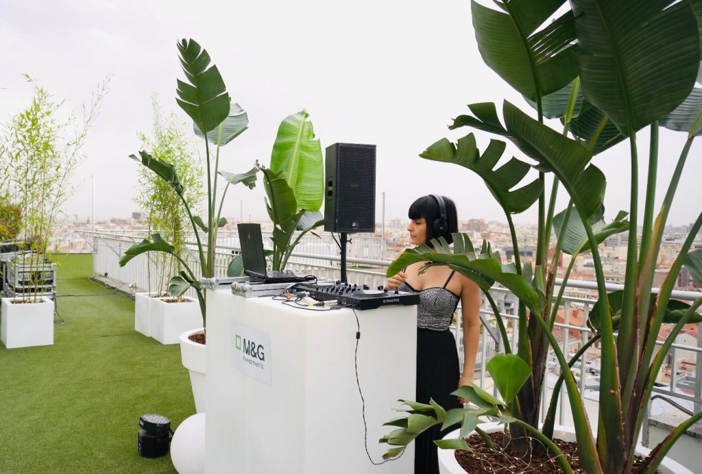 DJ Aleea tras una cabina blanca entre plantas durante un evento